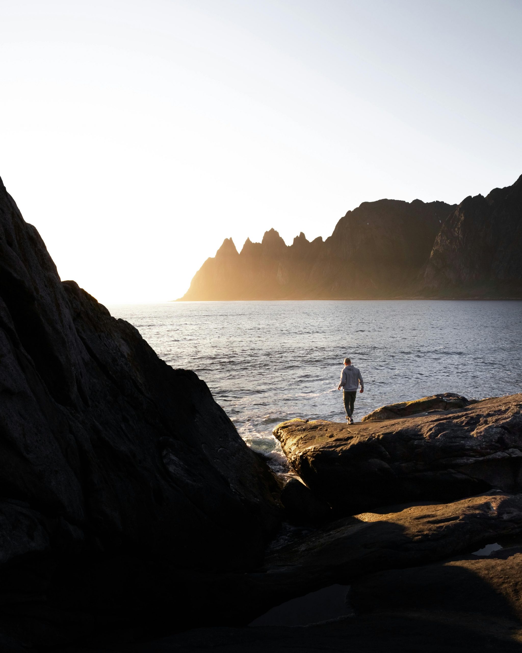 A solitary figure walks along a rocky shore with mountains and ocean in Norway during a stunning sunset.